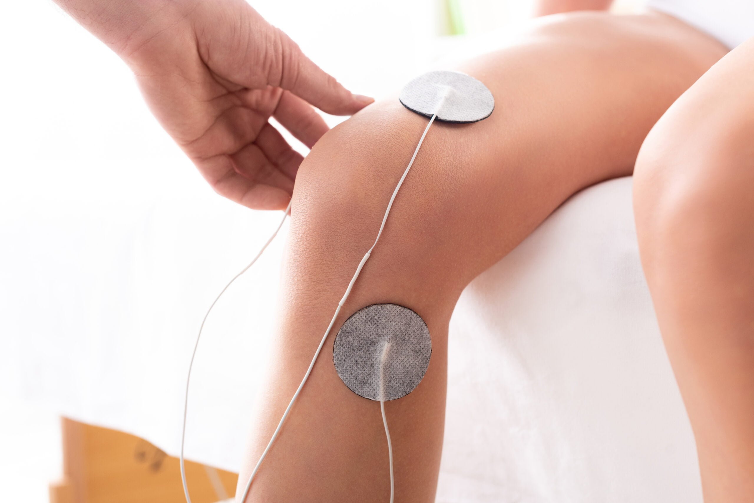 cropped-view-of-therapist-setting-electrodes-on-knee-of-patient-on-massage-couch-on-white-background